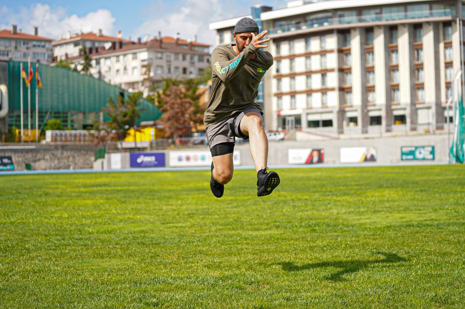 Energetic action photo of a man jumping on a grassy field in a cityscape background.