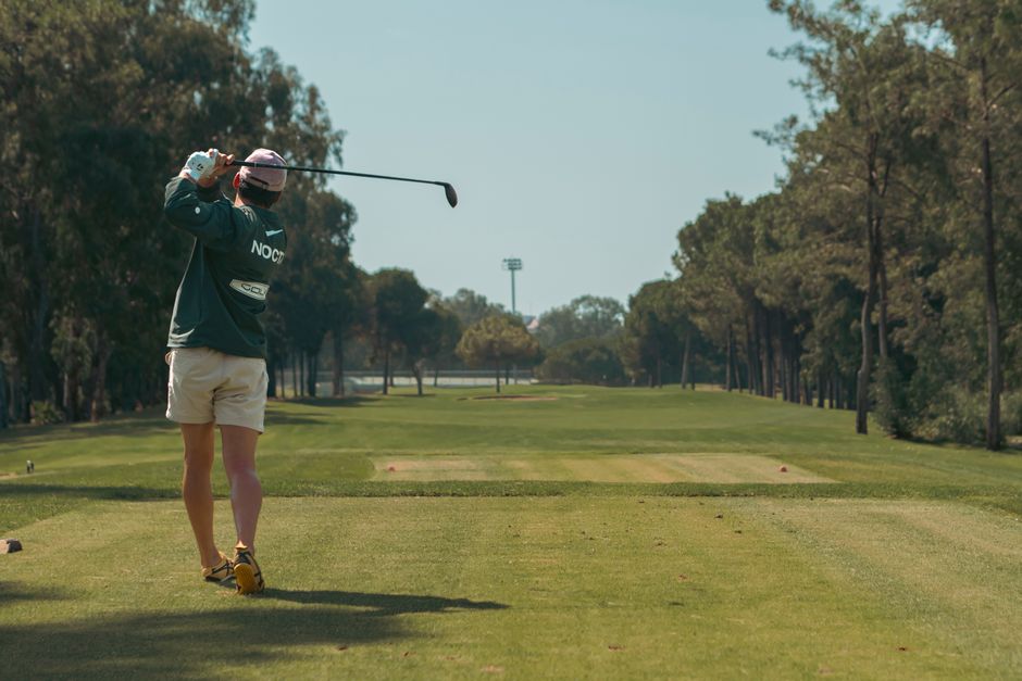 A golfer takes a powerful swing on a lush golf course surrounded by trees under a clear blue sky.