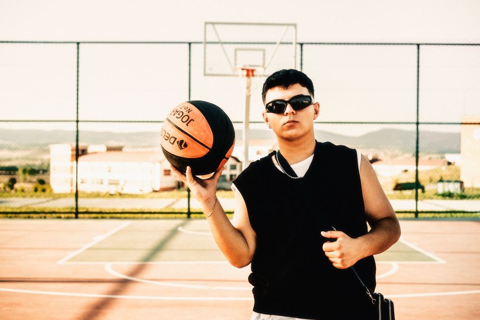 Confident young man in sunglasses holding a basketball at an outdoor court during the day.