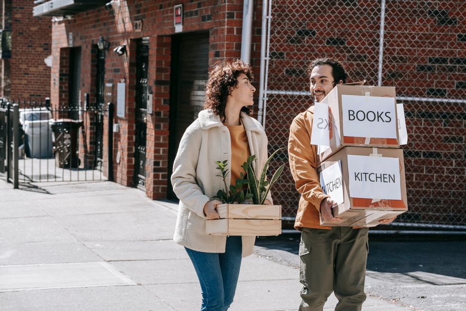 A couple happily moving to a new home carrying boxes and a plant on a sunny day.